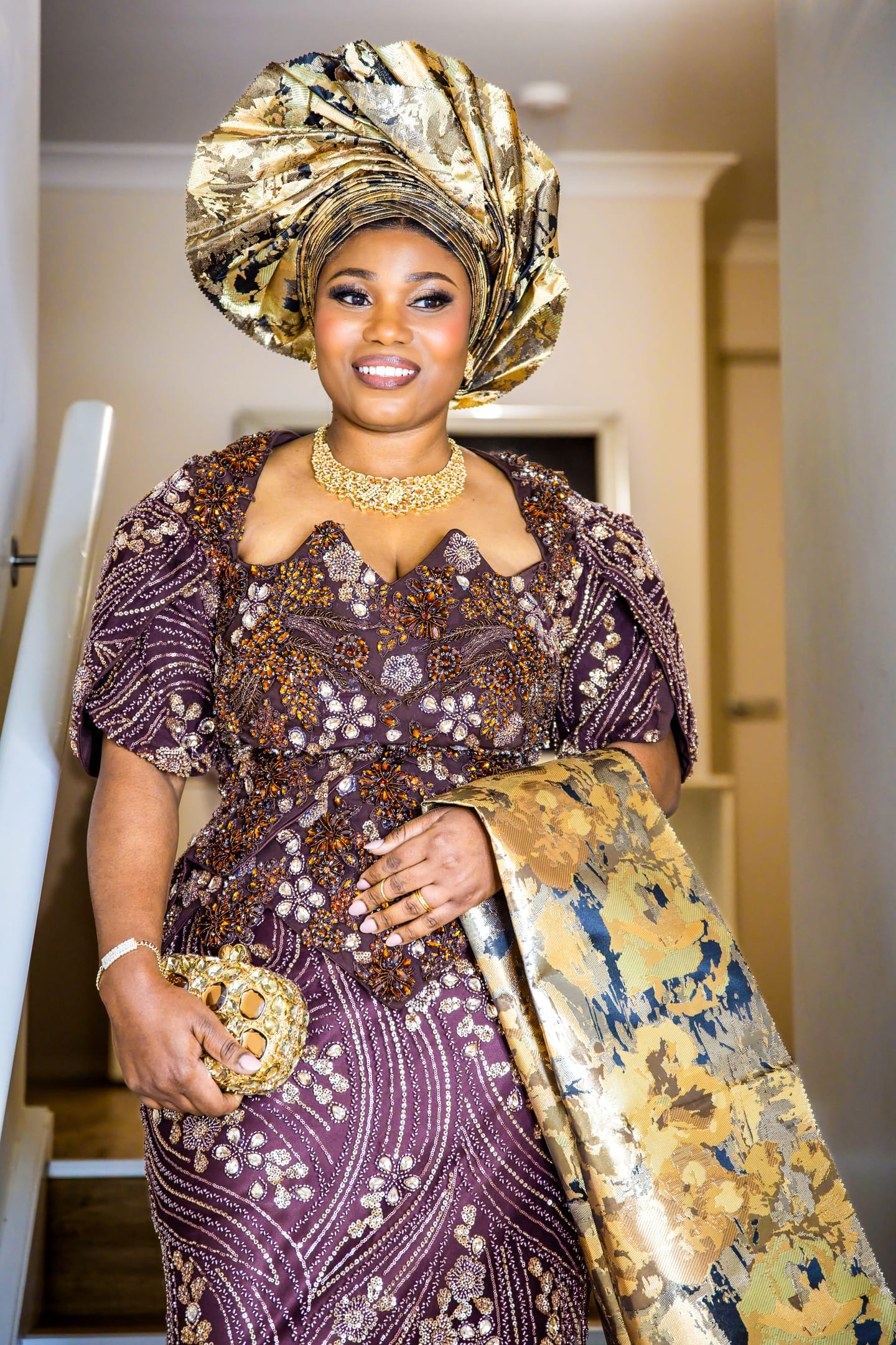 A woman on a staircase in an embellished brown and purple dress with a gold gele and choker
