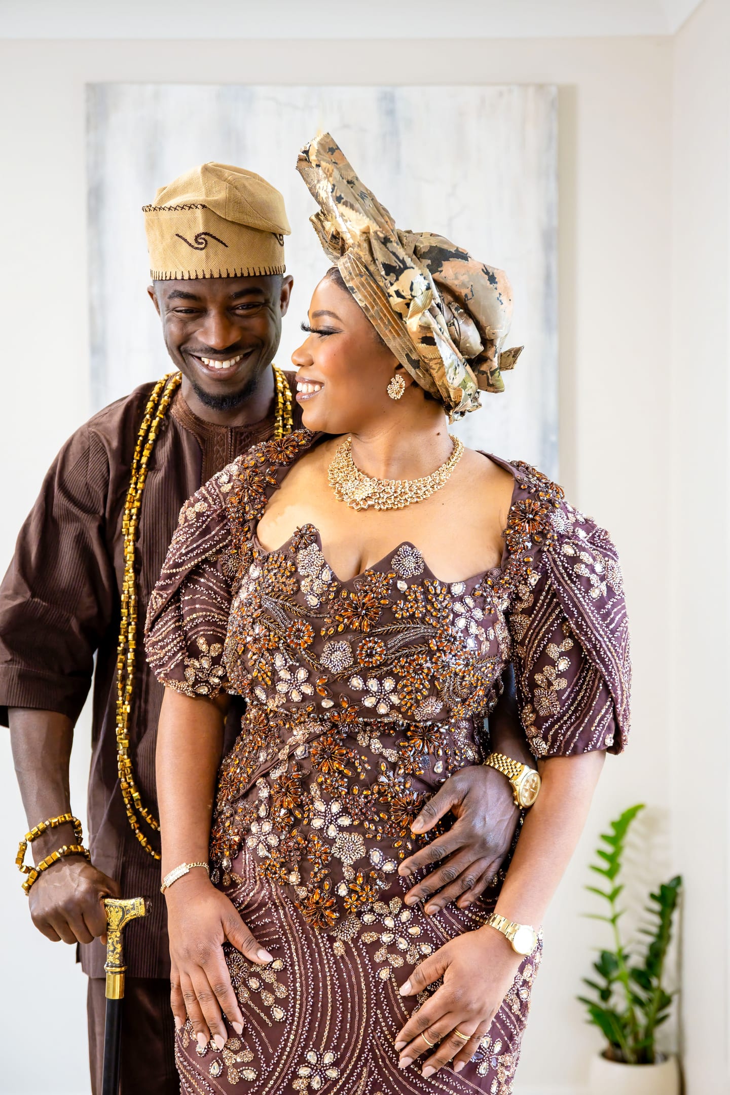 A couple standing close and laughing, he in a brown agbada with a walking stick, she in beaded brown lace