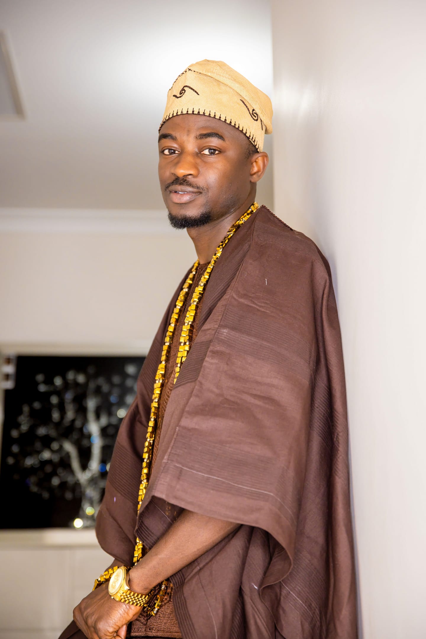 A man in a brown agbada and embroidered tan fila leaning against a white wall, gold beads at his neck