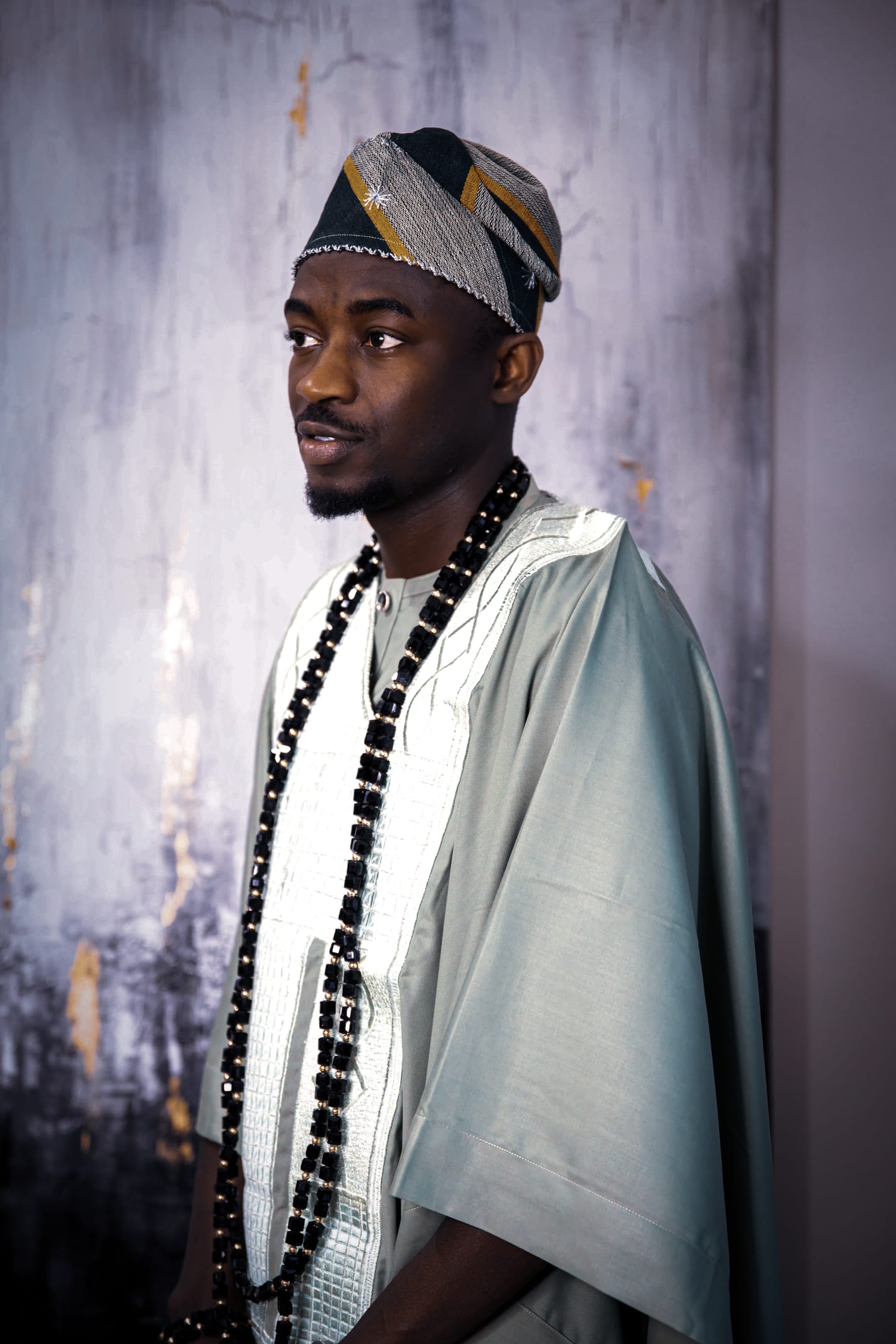 A man in a sage green agbada over white embroidery, black beads layered against a textured grey wall