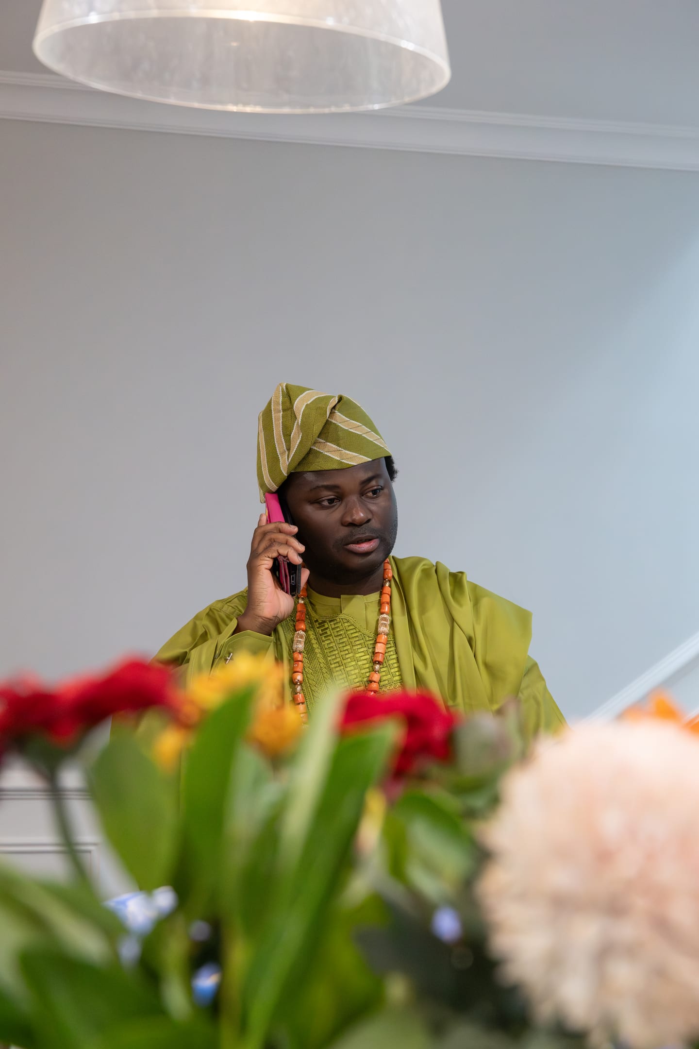 A guest in a green agbada and striped fila taking a phone call, framed by red and yellow flowers