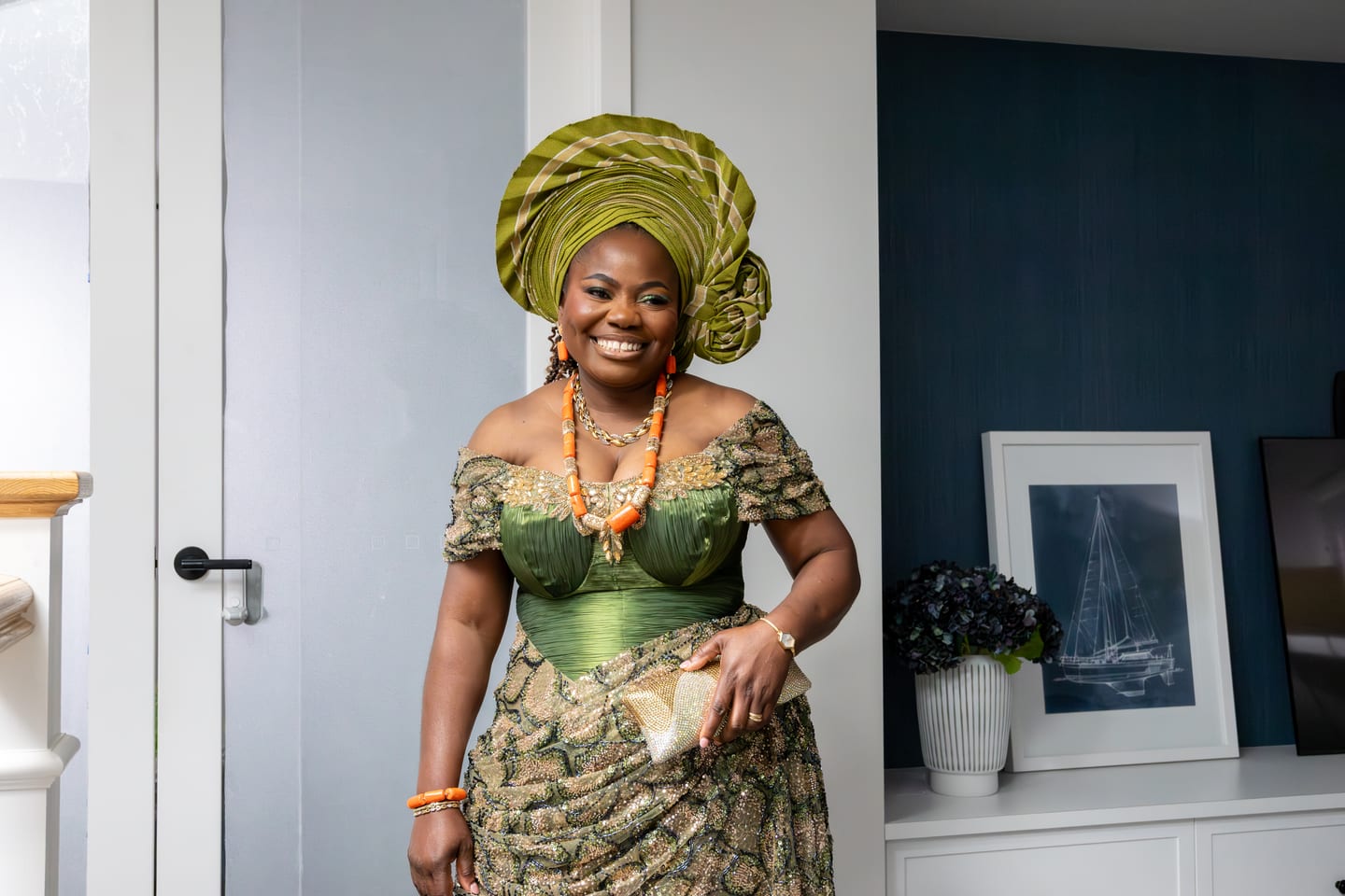 A woman in a green gele and beaded green dress laughing, wearing orange coral beads and holding a clutch
