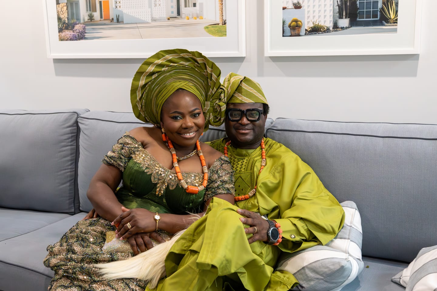 A couple in matching green and gold aso-ebi seated together on a grey sofa, both smiling at the camera