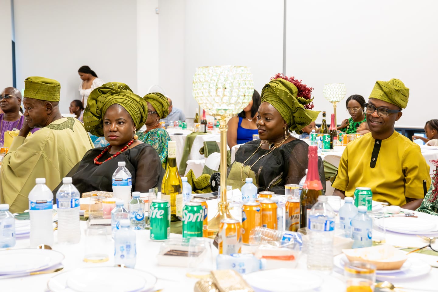 A long table of guests in green gele and gold buba, plates and drinks spread across the cloth