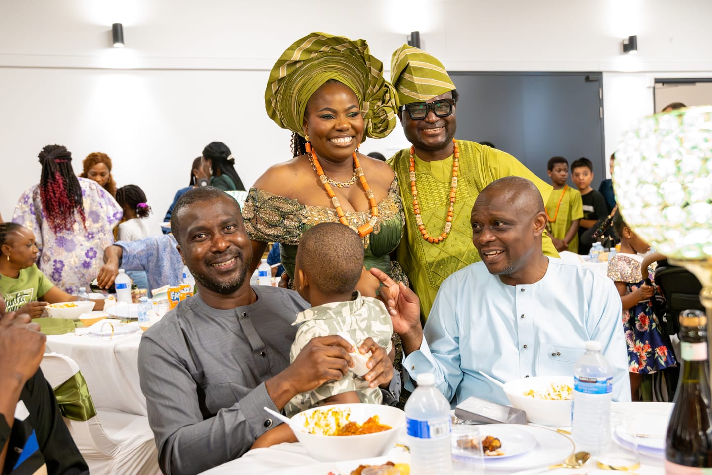 A family gathered around a table, a woman in a green gele standing behind two seated men and a small child
